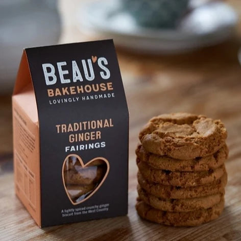 A package of Beau's Bakehouse Traditional Ginger Fairings cookies stands next to a stack of golden-brown ginger cookies on a wooden surface.